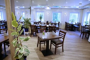 Un restaurante amplio con numerosas mesas y sillas, plantas en macetas y grandes ventanales que dejan entrar la luz natural.