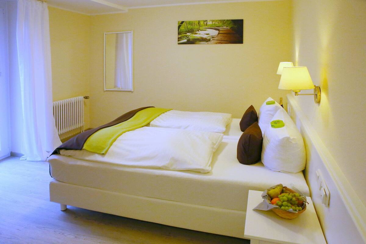 A neatly made bed with white linens and a green throw, a bedside table with a fruit basket, a wall-mounted lamp, and a painting of a forest scene.