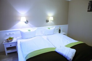 Quarto de hotel com cama bem arrumada, duas luminárias de cabeceira, uma pequena mesa de cabeceira com uma cesta de frutas e uma garrafa de champanhe.