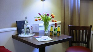 A hotel room with a small table featuring a bottle of Gerolsteiner, a glass of water, a glass of orange juice, and a vase with tulips. There is a chair and a magazine on the table. - Waldhotel Wilhelmshöhe (Freudenberg)