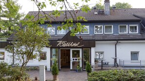 A two-story building with a sign reading "Taldhotel". There are potted plants in front of the entrance, a paved walkway, and a small outdoor seating area with tables and chairs. - Waldhotel Wilhelmshöhe (Freudenberg)