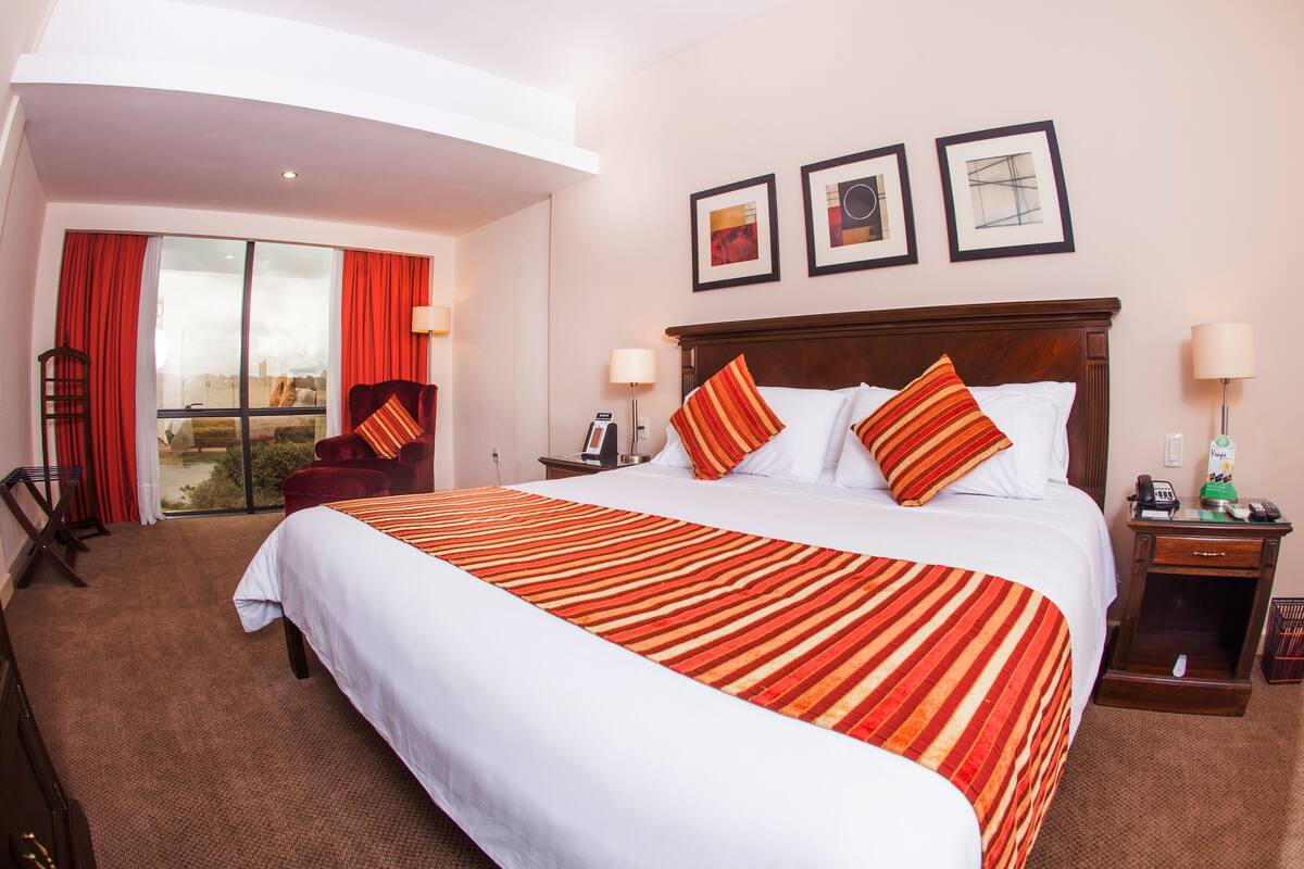 A hotel room with a large bed, a desk with a lamp, a chair, a window with curtains, and artwork on the wall.