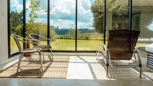 A room with large windows offering a view of a green landscape and a city skyline. There are two wicker chairs and a small table. - Hotel Casino Acaray (Ciudad Del Este)