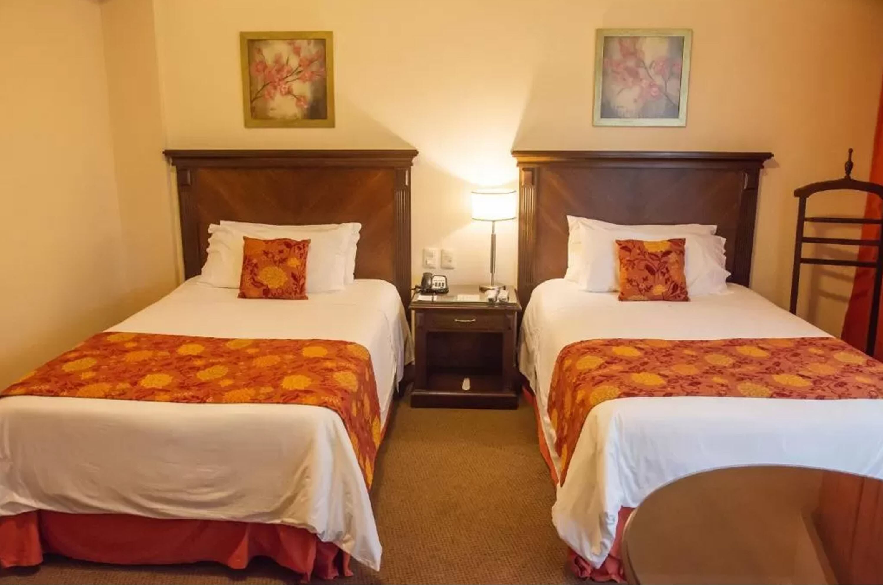 Ein Hotelzimmer mit zwei Betten, beide mit einem hölzernen Kopfteil, weißer Bettwäsche und orangefarbenen Blumendecken. Zwischen den Betten steht ein Nachttisch mit einer Lampe, und in der Ecke ist ein Sessel zu sehen.