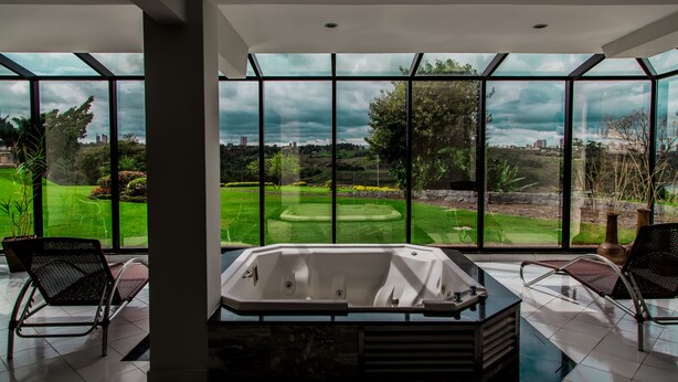 A modern bathroom with a large bathtub, two chairs, and a view of a landscaped garden through large windows. - Hotel Casino Acaray (Ciudad Del Este)