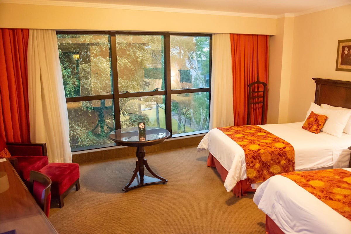 A hotel room with two beds, a small table, and a view of trees through large windows.