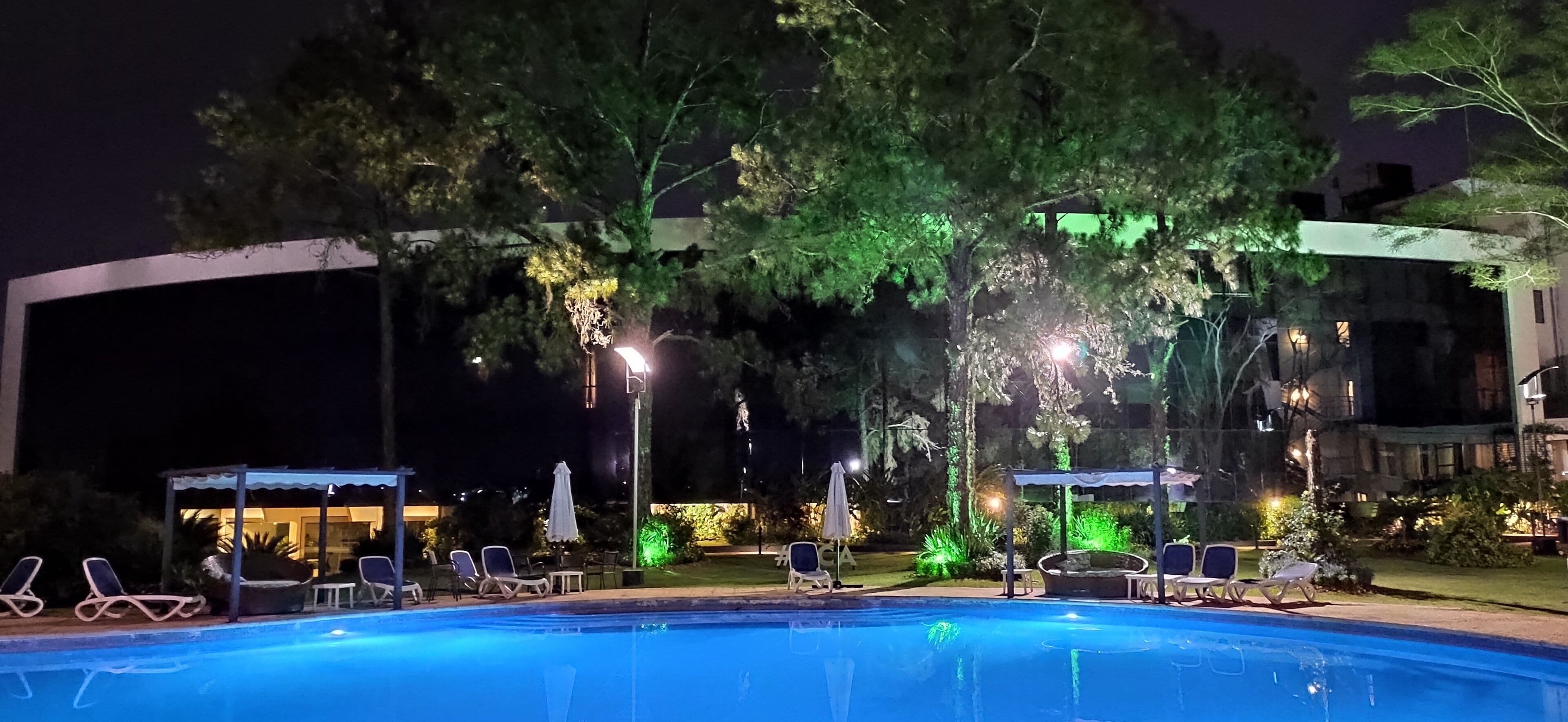 Zona de piscina nocturna con sillones reclinables y sombrillas, rodeada de árboles y edificios.