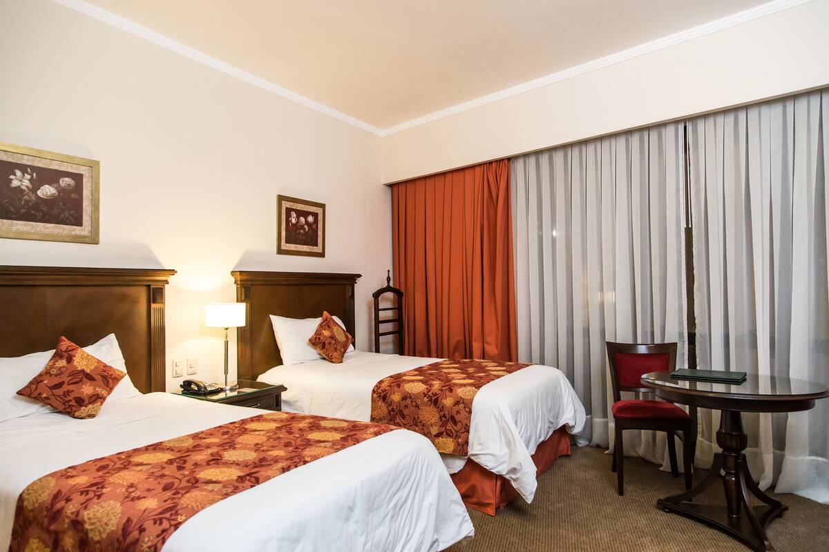A hotel room with two beds, a desk, a chair, a table, and curtains.
