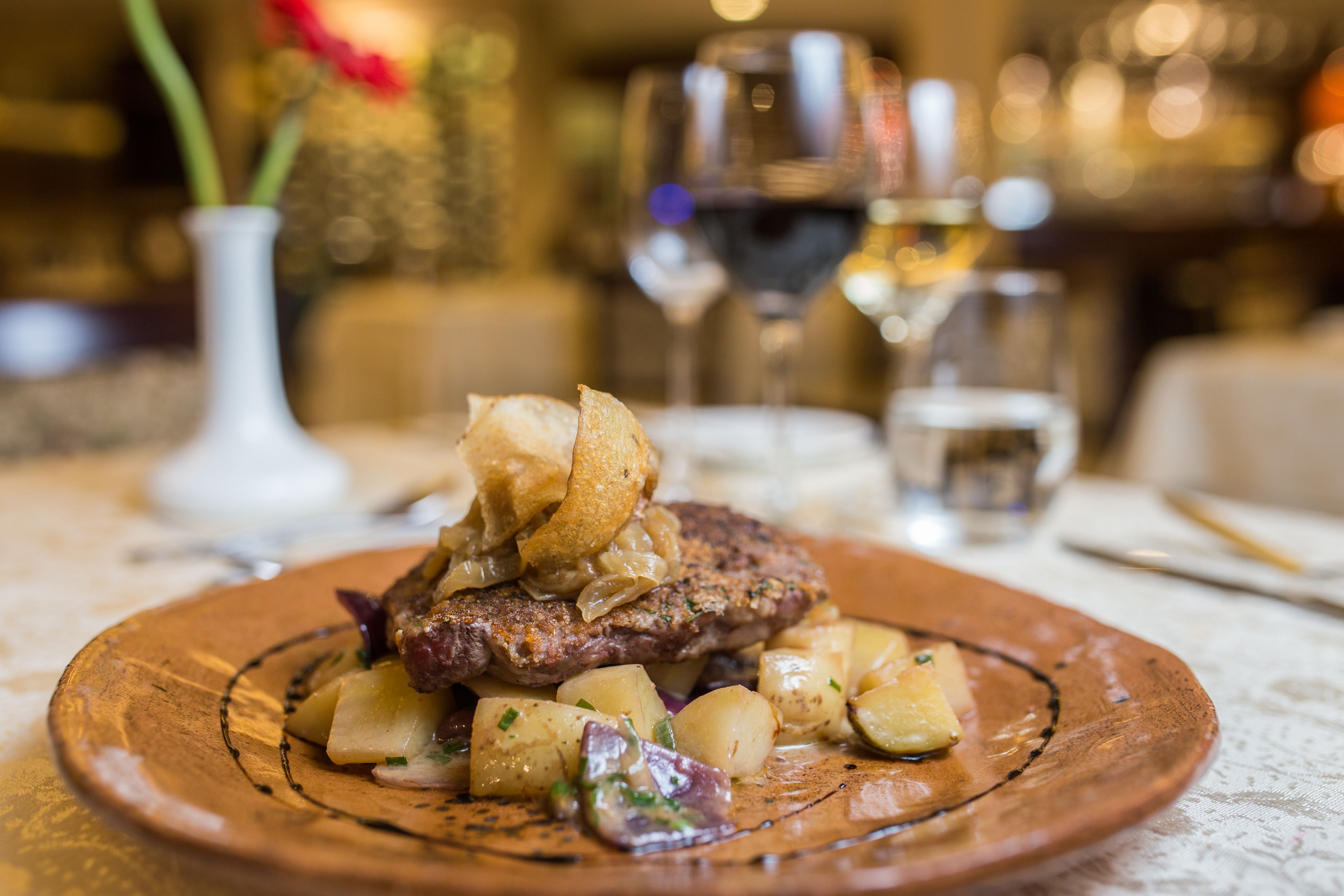A plate of food with a steak, potatoes, and onions. There are glasses of wine and a vase with flowers in the background.
