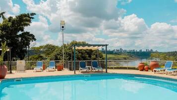 A pool area with lounge chairs and a pergola, overlooking a city skyline and a river.