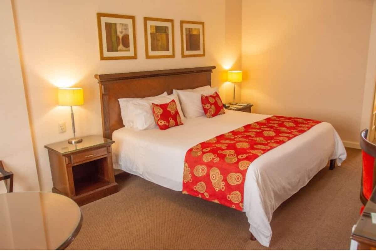 A hotel room with a wooden bed, white bedding, red and gold patterned bedspread, two bedside lamps, and a small wooden nightstand.