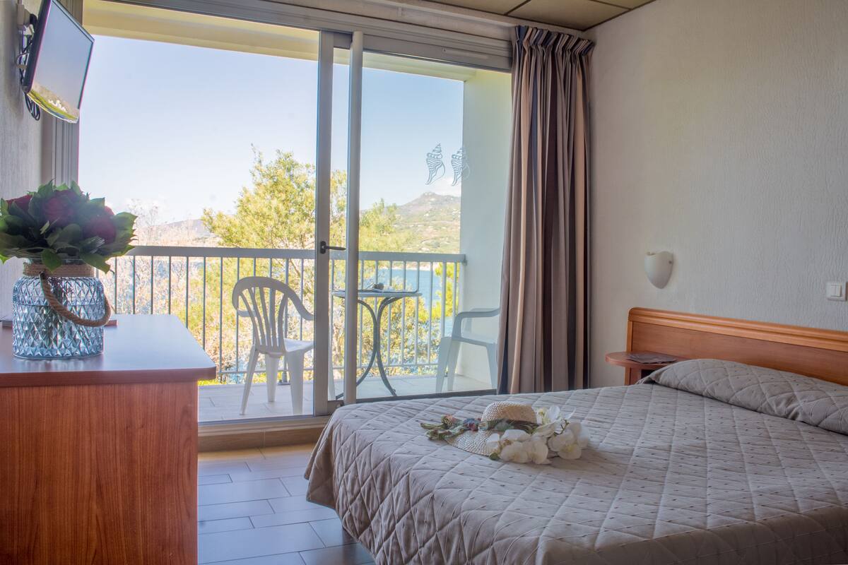 Habitación de hotel con una cama, un escritorio con un florero, una silla, una mesa y vistas al exterior.