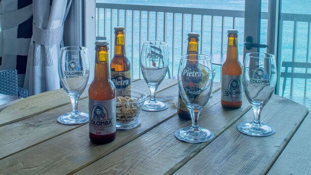 A wooden table with four glasses and two bottles of beer on a balcony overlooking the sea. - Hôtel Roc e Mare (Propriano)