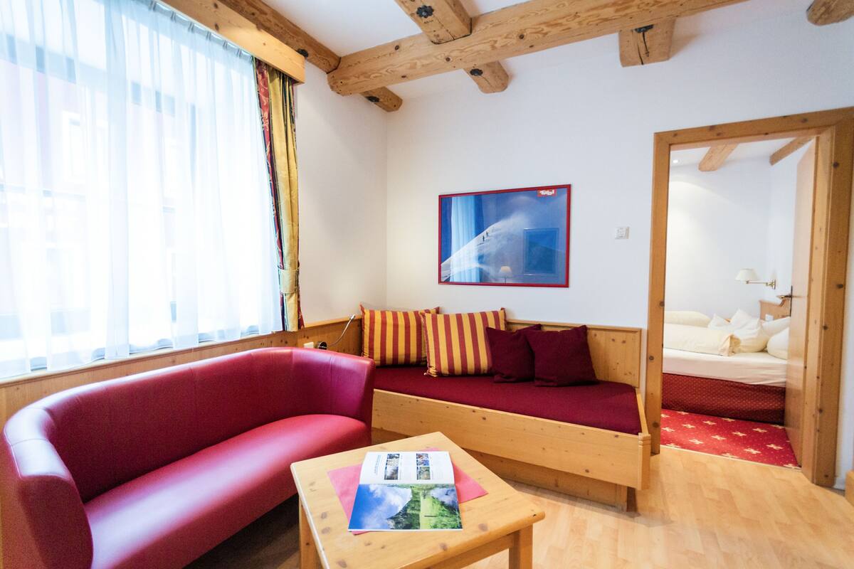 Een knus ogende kamer met een rode bank, een houten tafel met een tijdschrift en een bed met een rood-witte dekbedovertrek.