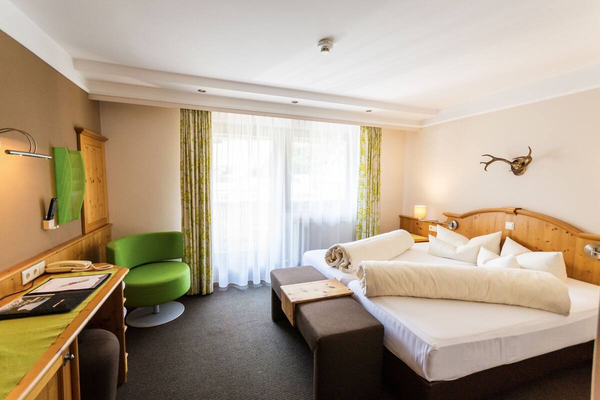 Hotelkamer met een bed, een bureau, een groene stoel, een houten hoofdeinde en een decoratieve geweivleugel aan de muur.