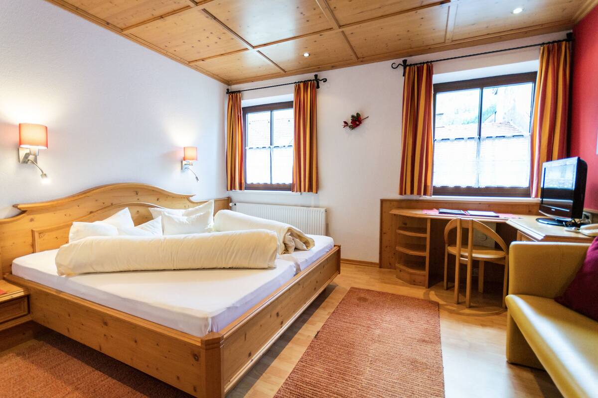 Een houten bed met witte lakens, een bureau met stoel, een televisie en een raam met gordijnen.