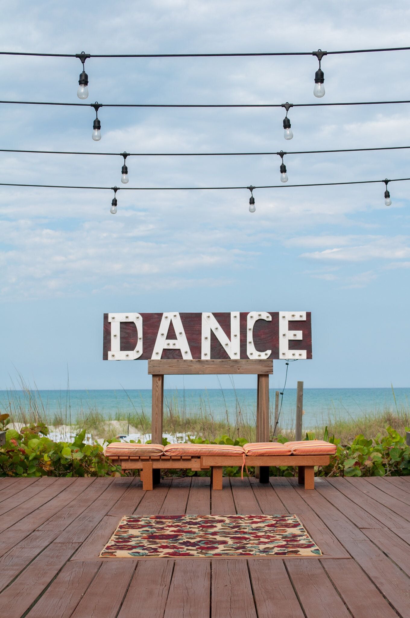 A wooden deck with a bench and a rug, a sign that says "DANCE", and a string of lights against a beach backdrop.