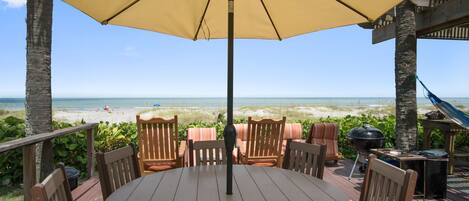 A deck with a round table and chairs, overlooking a beach.