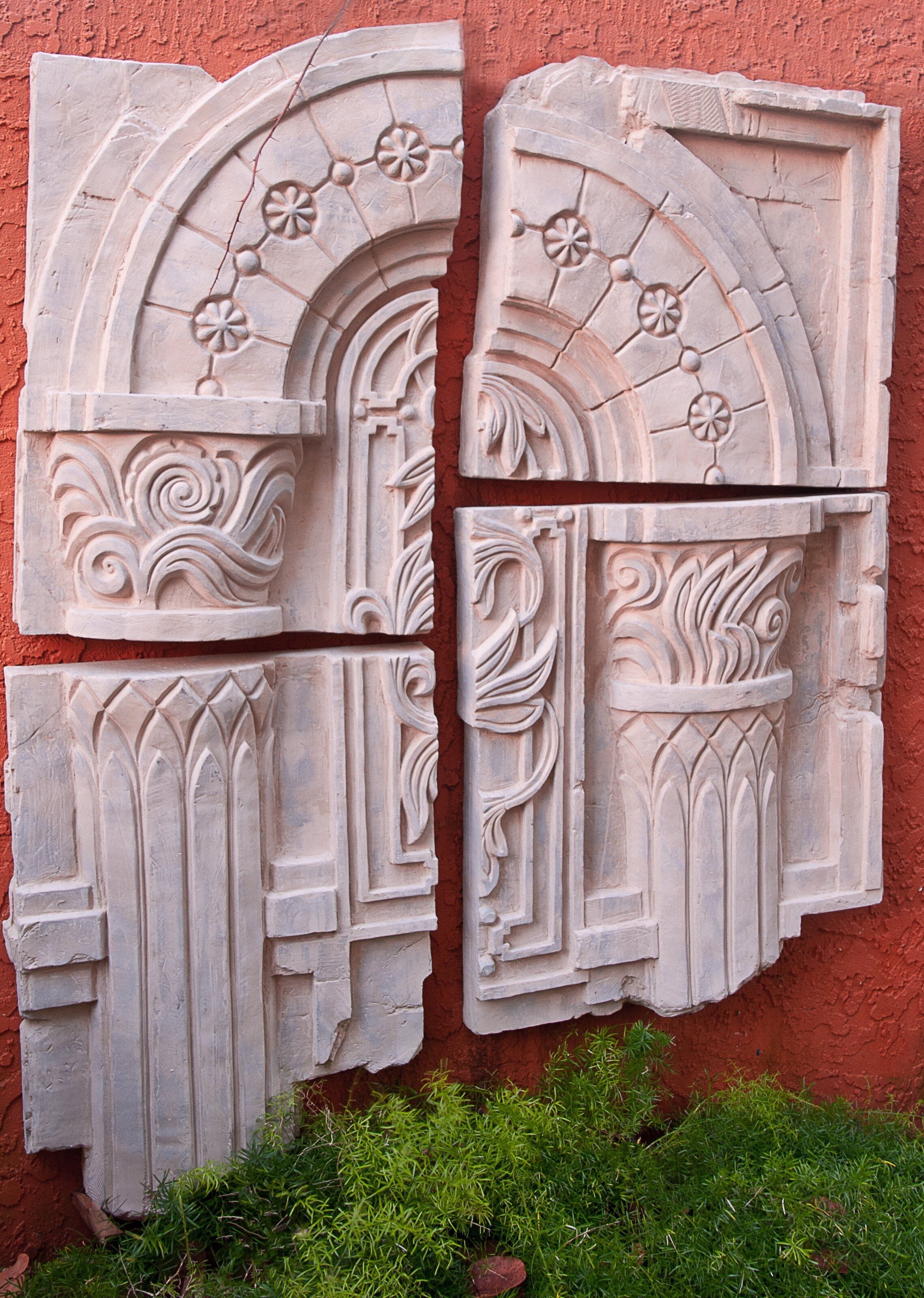 Un élément architectural en pierre, orné de motifs floraux et de volutes sculptés, qui se détache sur un mur rouge.