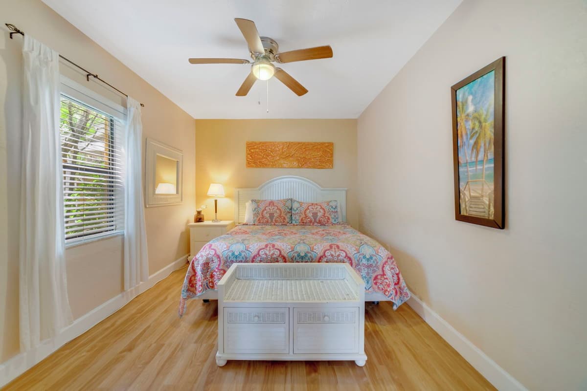 A bedroom with a bed, ceiling fan, bedside lamps, a painting, and a window with blinds.