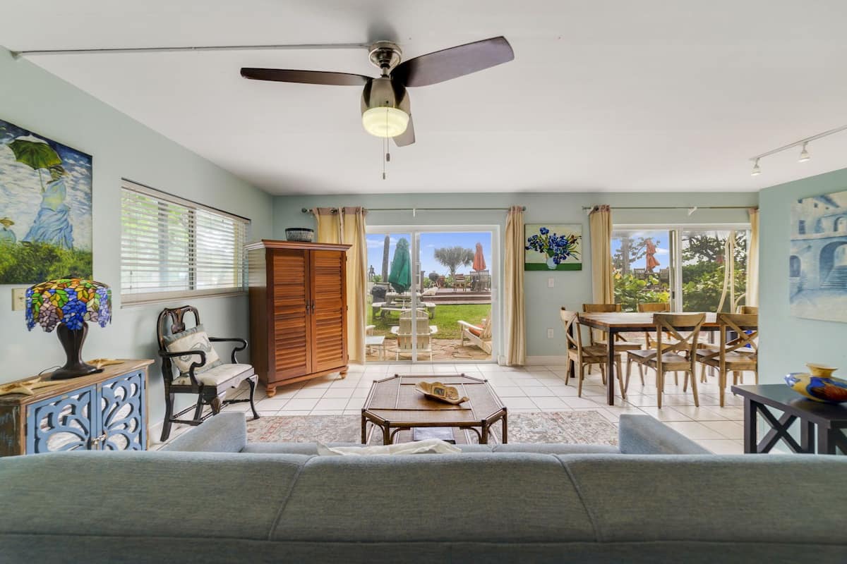 A spacious living area with a ceiling fan, a dining table with chairs, a sofa, and a view of the outdoors through large windows.