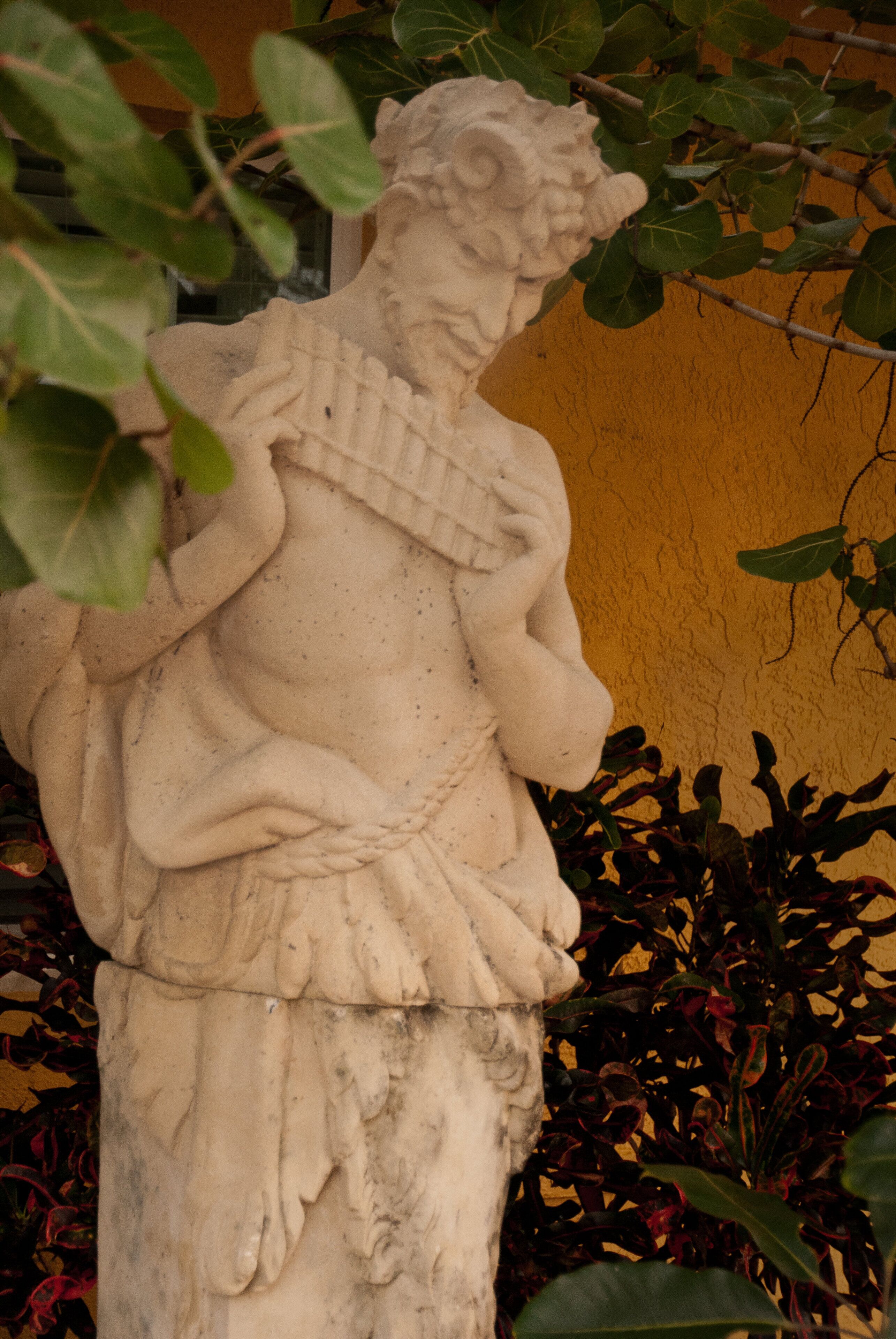A stone statue of a figure with a detailed robe, set against a yellow wall, surrounded by foliage.