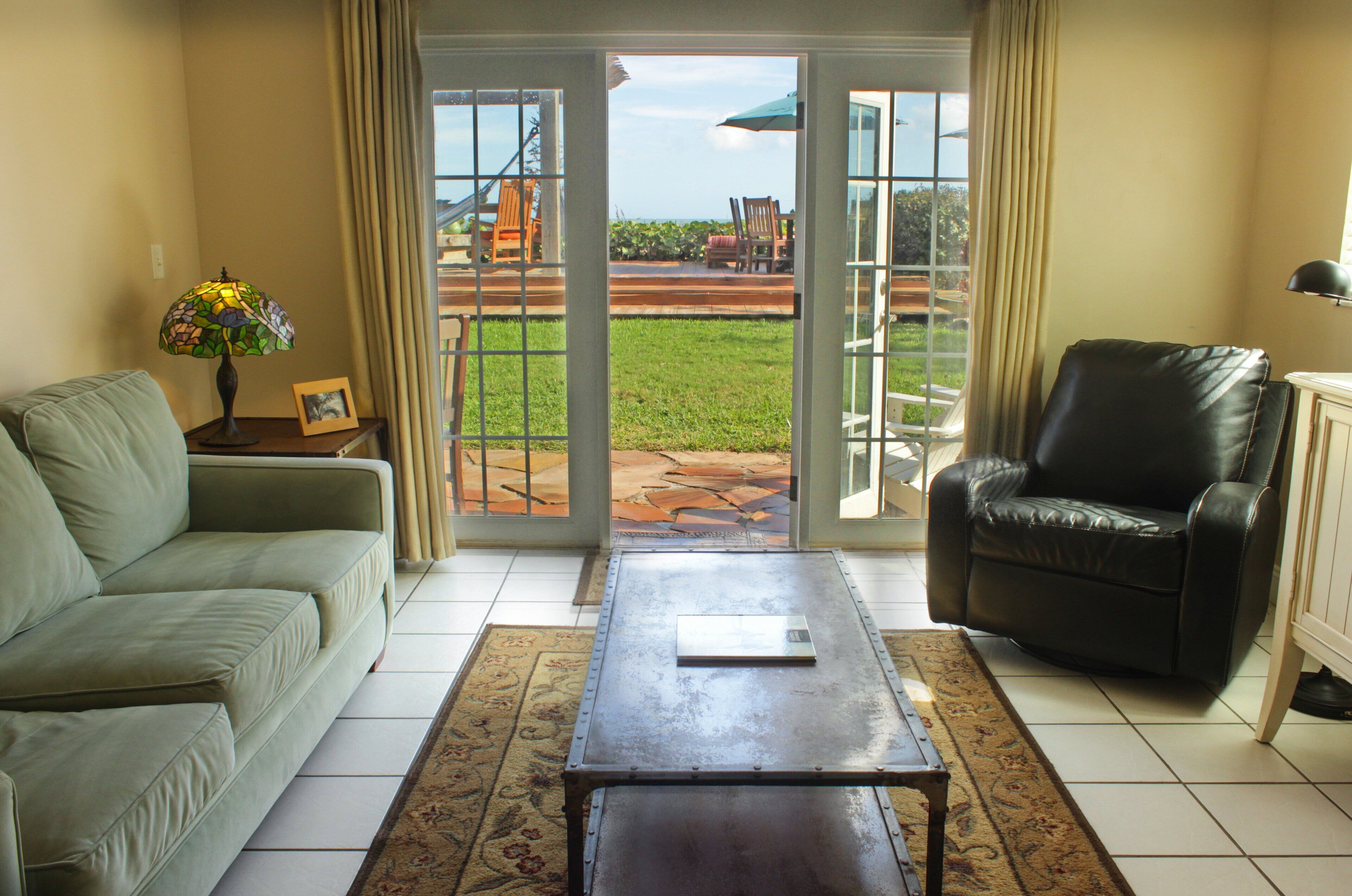 A living room with a sofa, a coffee table, a leather chair, and a view through glass doors to an outdoor patio area.