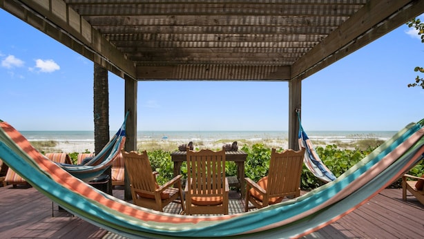 A wooden deck with a hammock, chairs, and a table overlooking the beach.