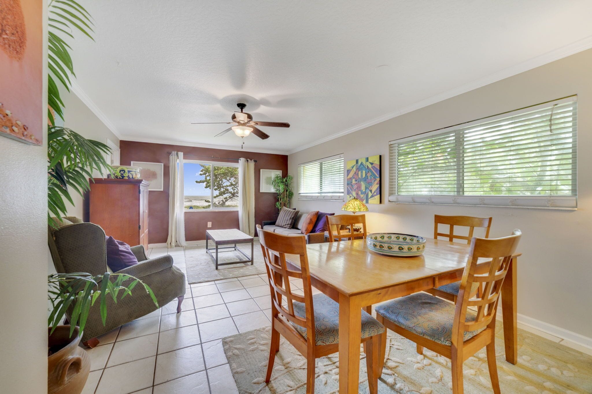 A dining area with a wooden table and chairs, a ceiling fan, and a view of the outdoors through large windows.