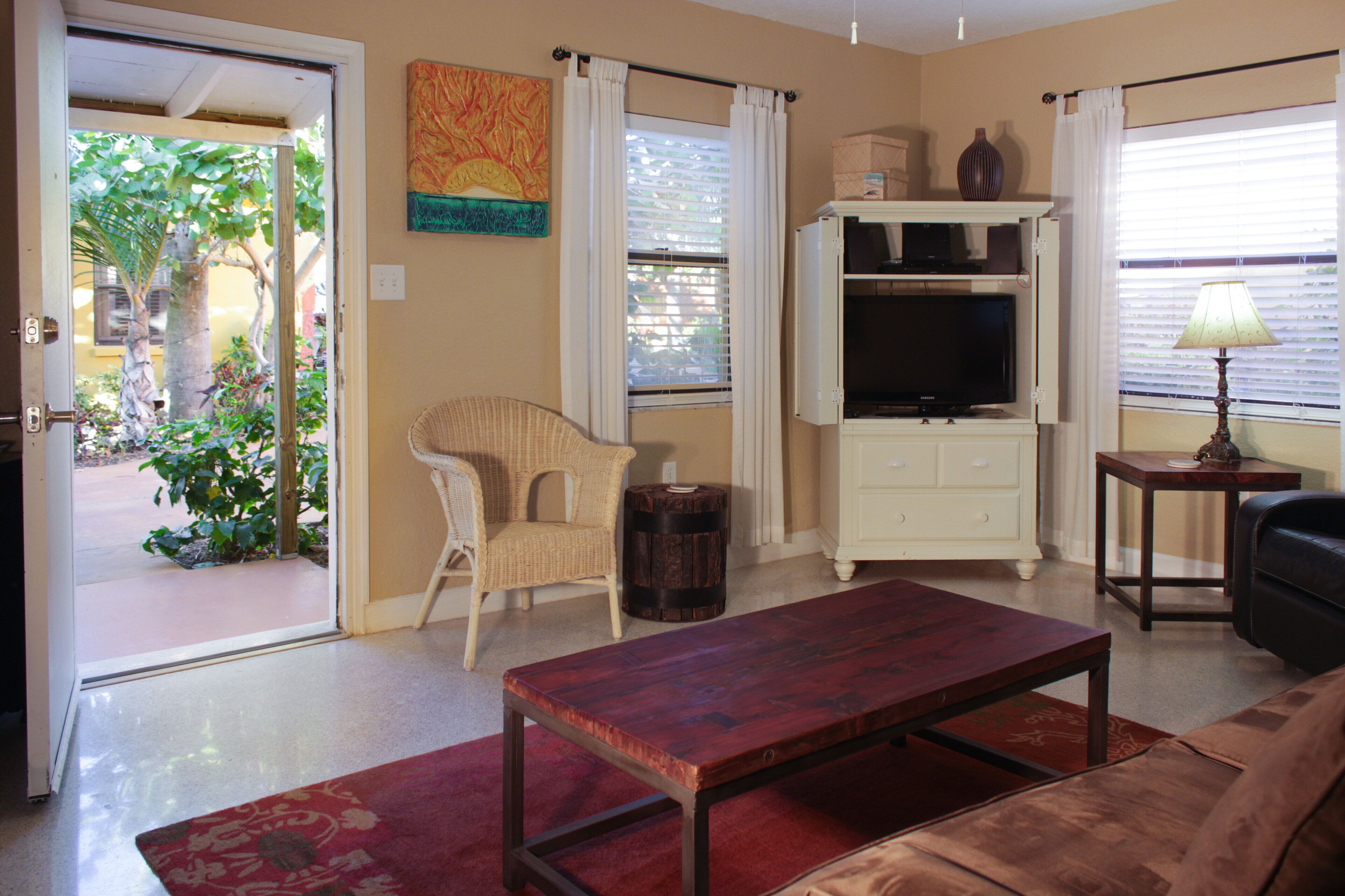 A living room with a television, a sofa, a coffee table, and a chair. There is a door leading to an outdoor area with plants.