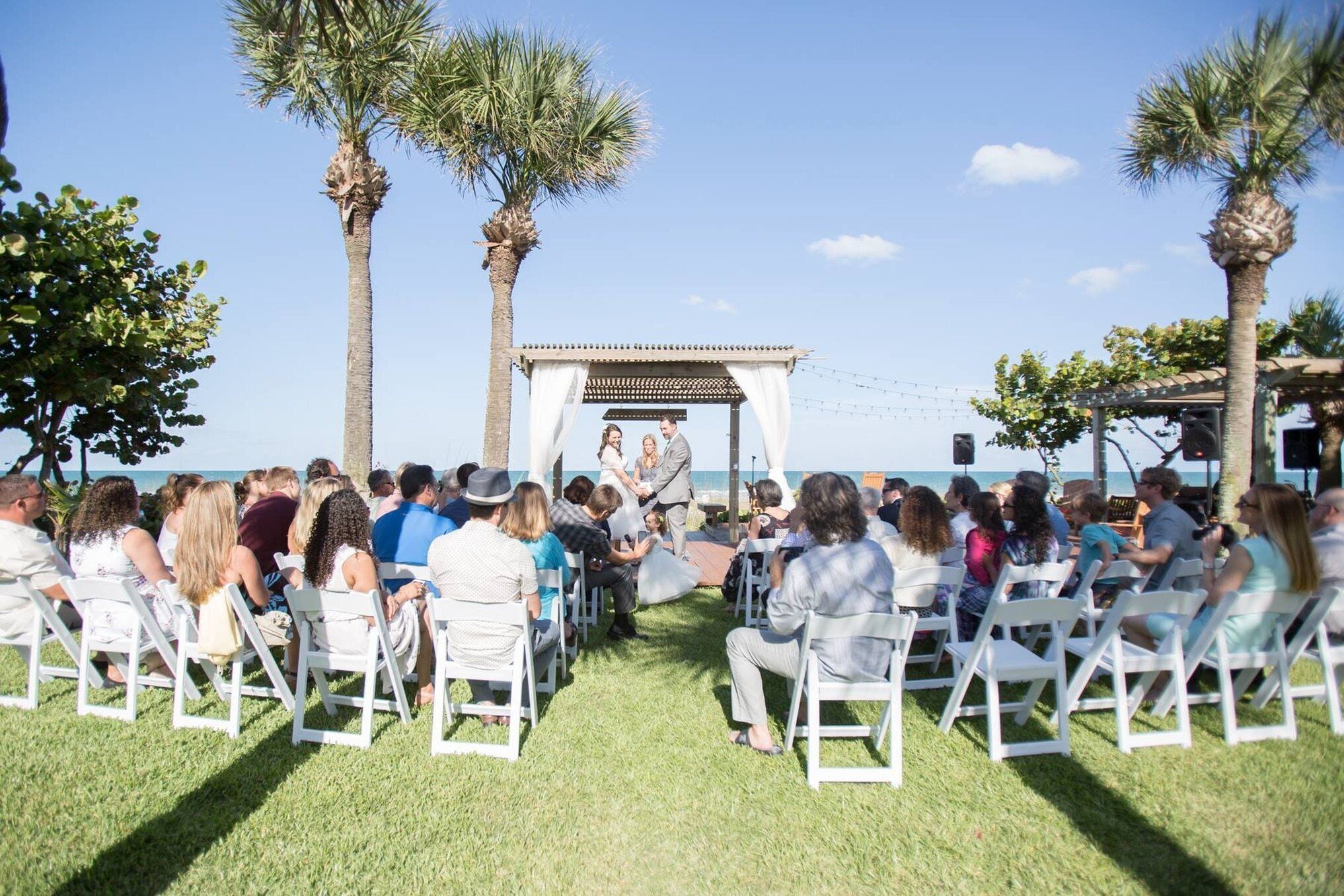 An outdoor wedding ceremony with guests seated on white chairs facing a couple under a pergola.