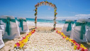 A beach wedding arch with white chairs and teal sashes, surrounded by white flowers.