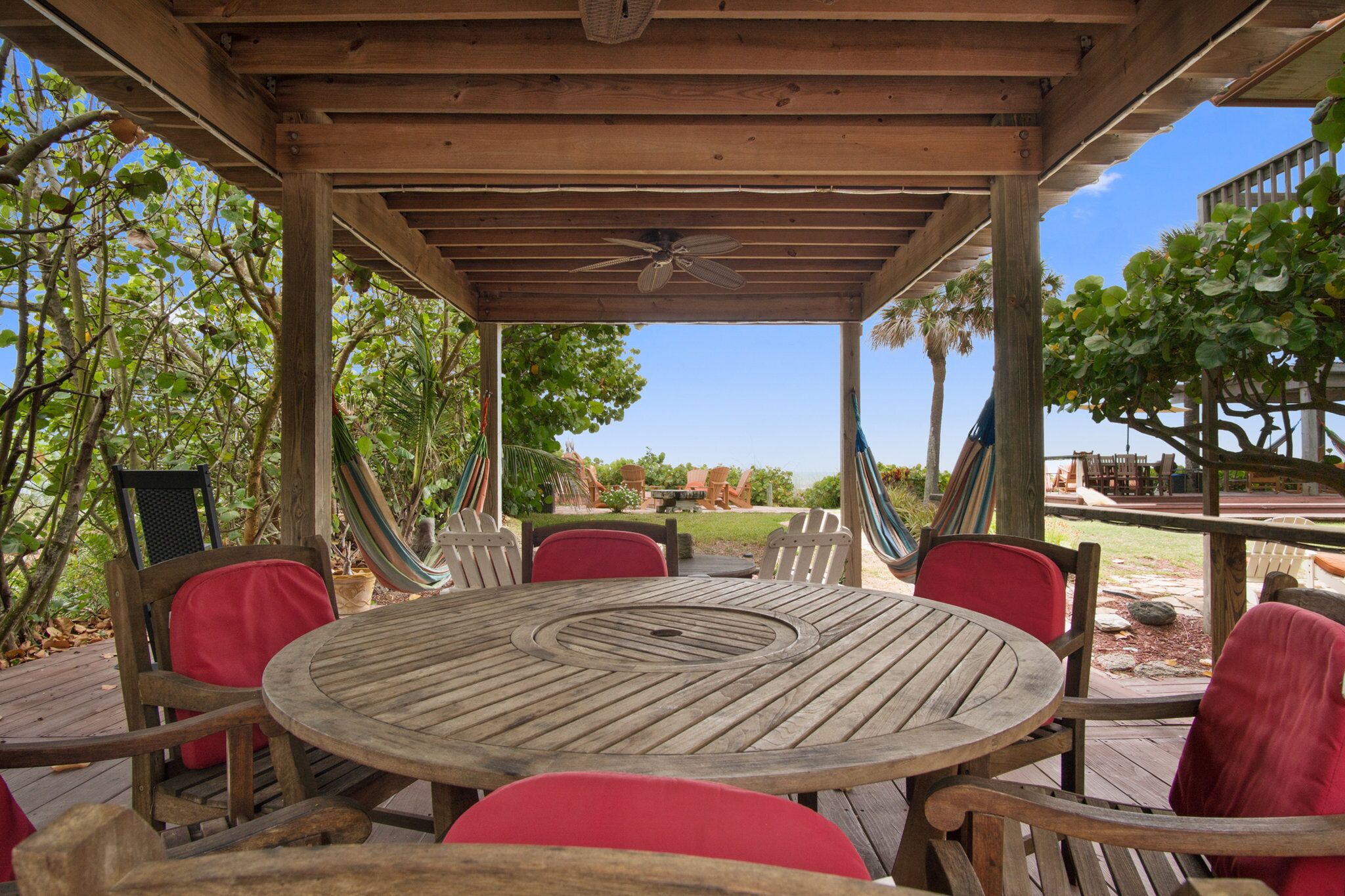 Eine Holzterrasse mit rundem Tisch und roten Stühlen, umgeben von Bäumen und Blick auf den Ozean.