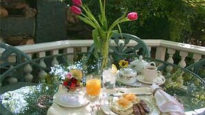 Une table ronde en verre, mise en place pour le petit-déjeuner, avec une théière, des tasses, une assiette de nourriture et un vase rempli de fleurs.