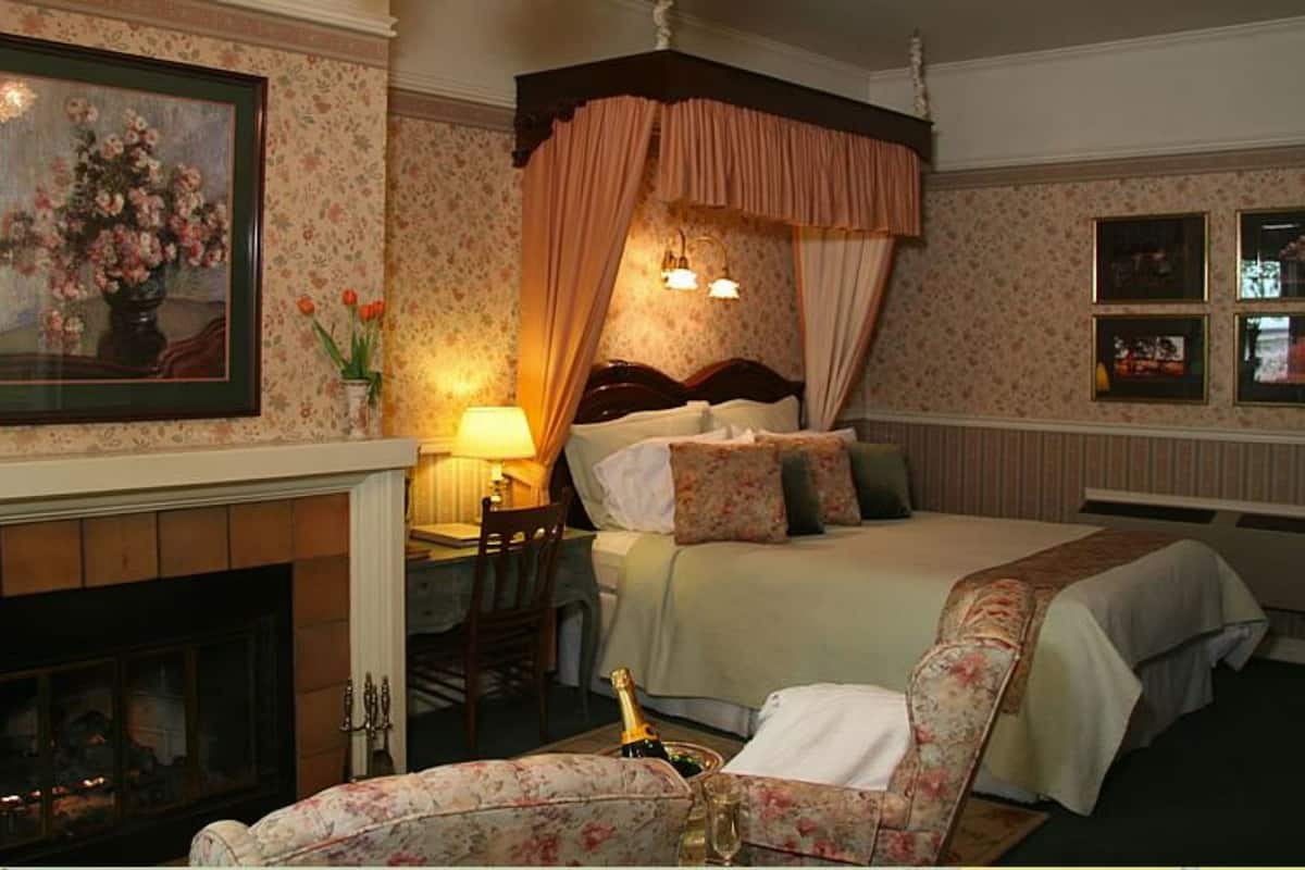 Ein Hotelzimmer mit Kamin, einem Himmelsbett, einem Schreibtisch mit Lampe und gerahmten Bildern an der Wand.
