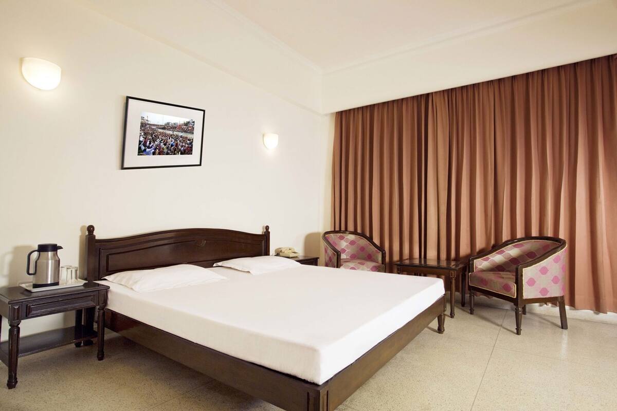 Una habitación de hotel con una cama grande, una mesita de noche con cafetera, dos sillas, un cuadro en la pared y una cortina.