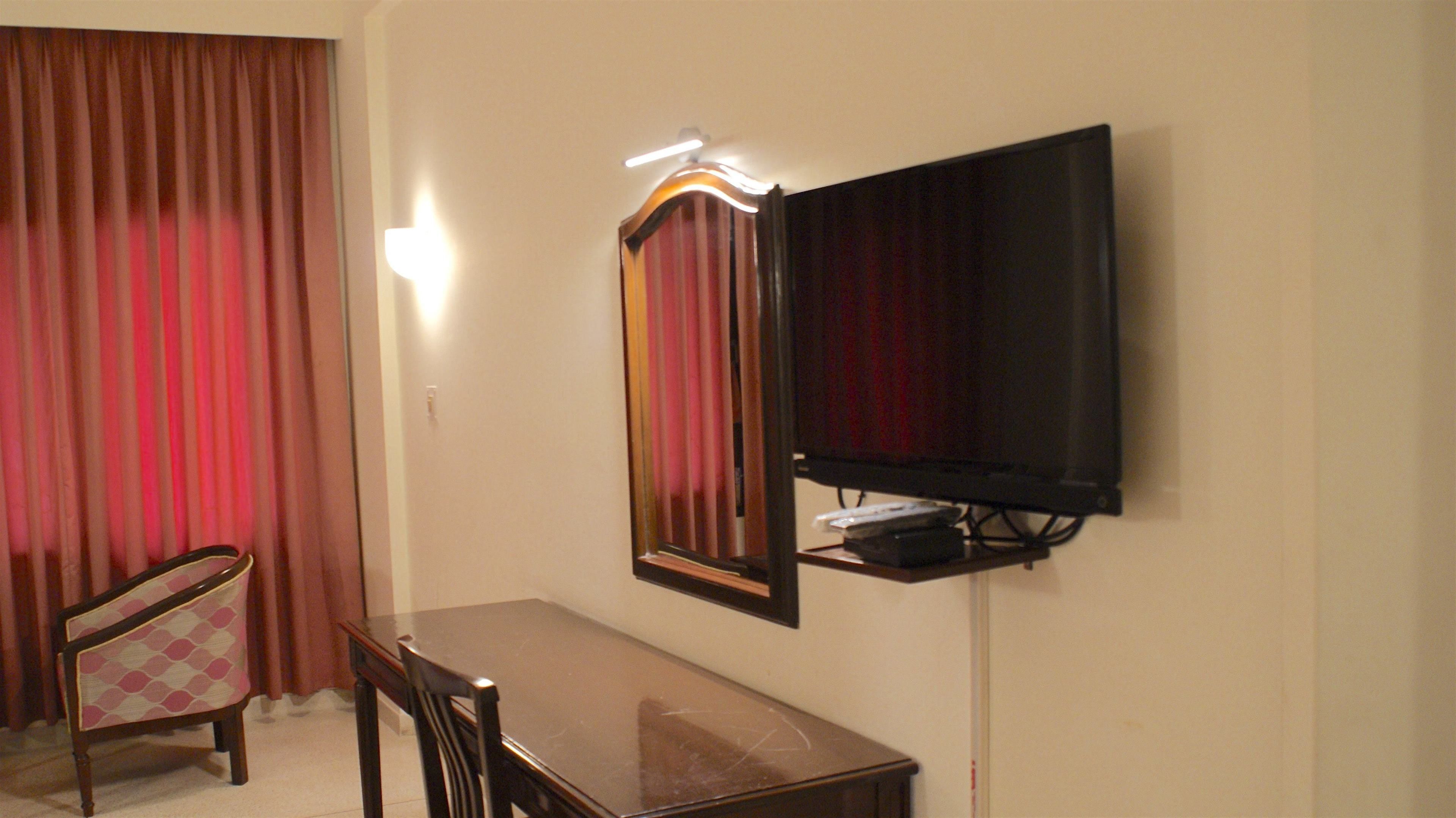 A room with a TV mounted on the wall, a wooden table, a chair, and a window with red curtains.