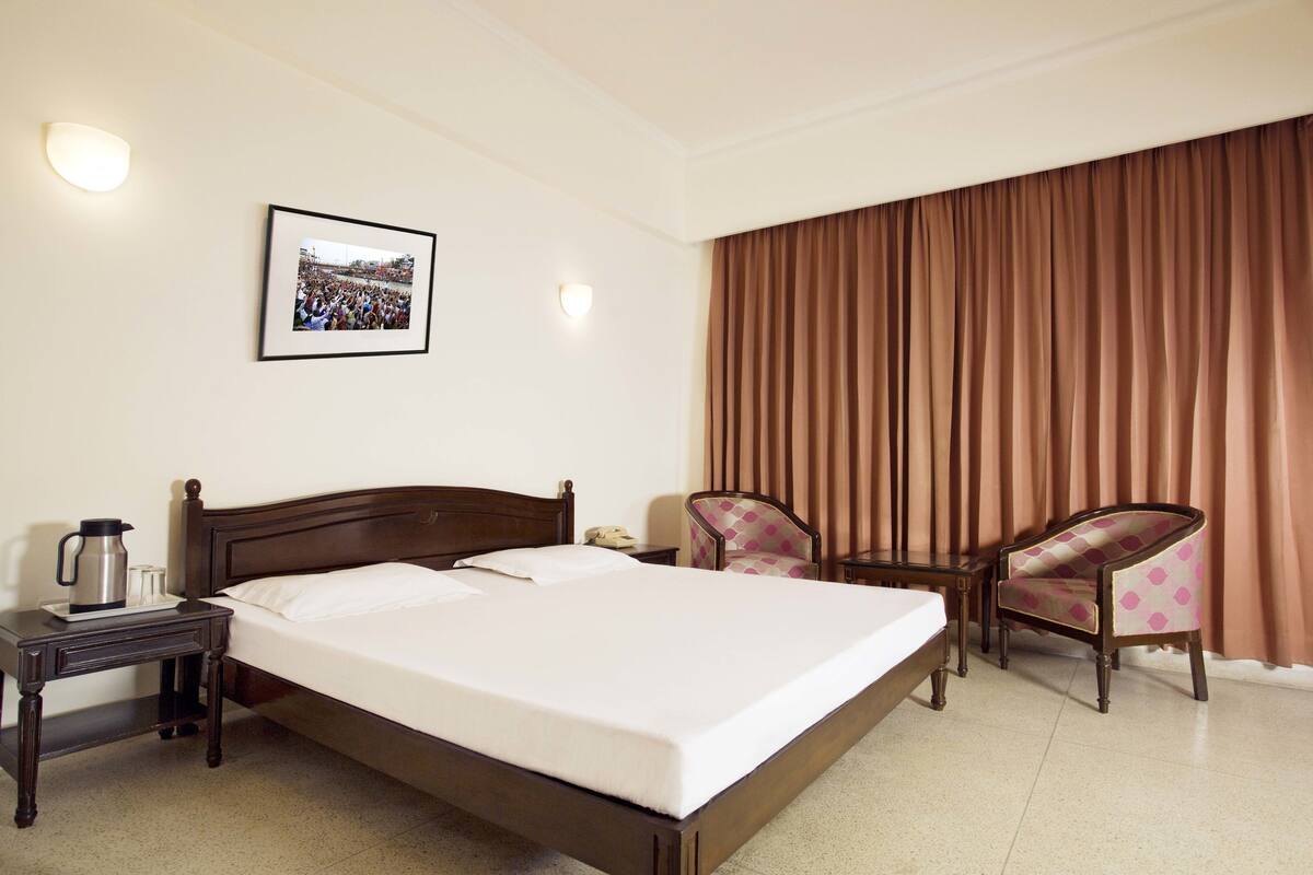 Habitación de hotel con una cama grande, una mesita de noche con una cafetera, dos sillas y un cuadro en la pared.