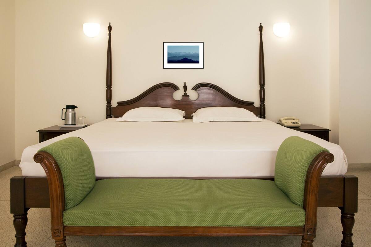 Una cama con dosel, con un banco tapizado de color verde, una mesita de noche con lámpara y un cuadro enmarcado en la pared.