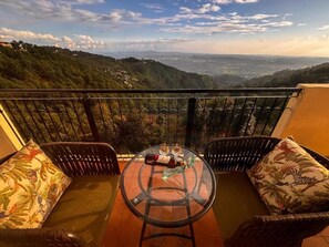 En balkon med fletmøbler og et glasbord, med udsigt over et bjergrigt landskab.