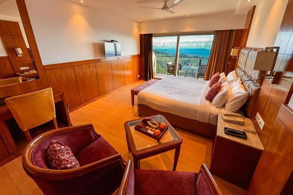 A hotel room with a large bed, a desk, a chair, a small table with fruit, a TV, and a balcony with a view.