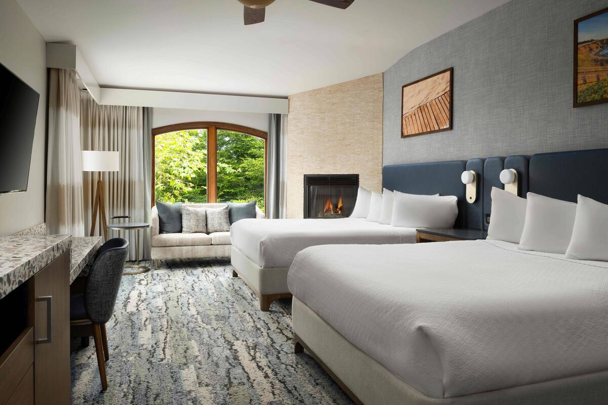 A hotel room with two beds, a desk, a chair, a fireplace, and a large window with a view of greenery.