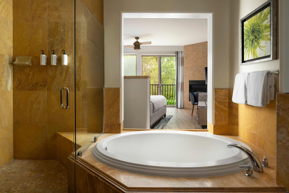 A modern bathroom with a large oval bathtub, a glass-enclosed shower, and a view of a bedroom with a balcony.