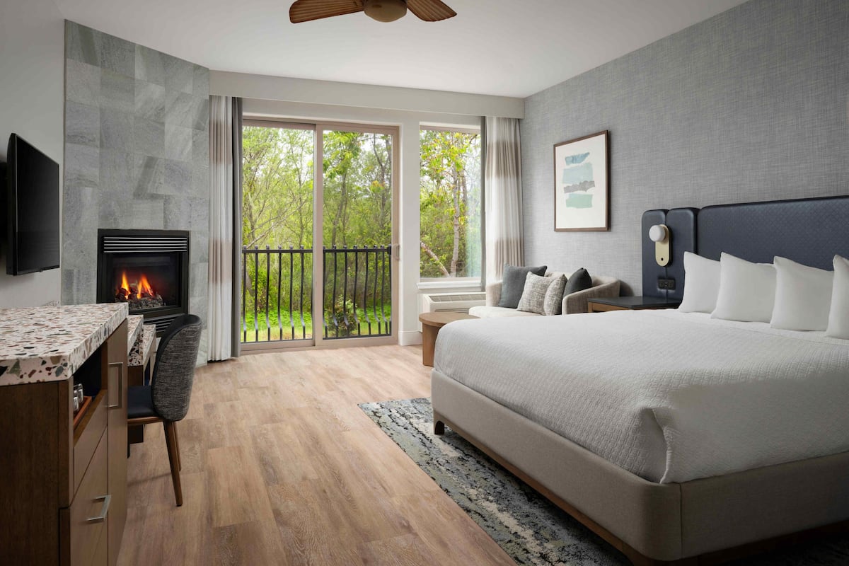 A modern bedroom with a large bed, a desk with a fireplace, a TV, and a balcony with a view of trees.