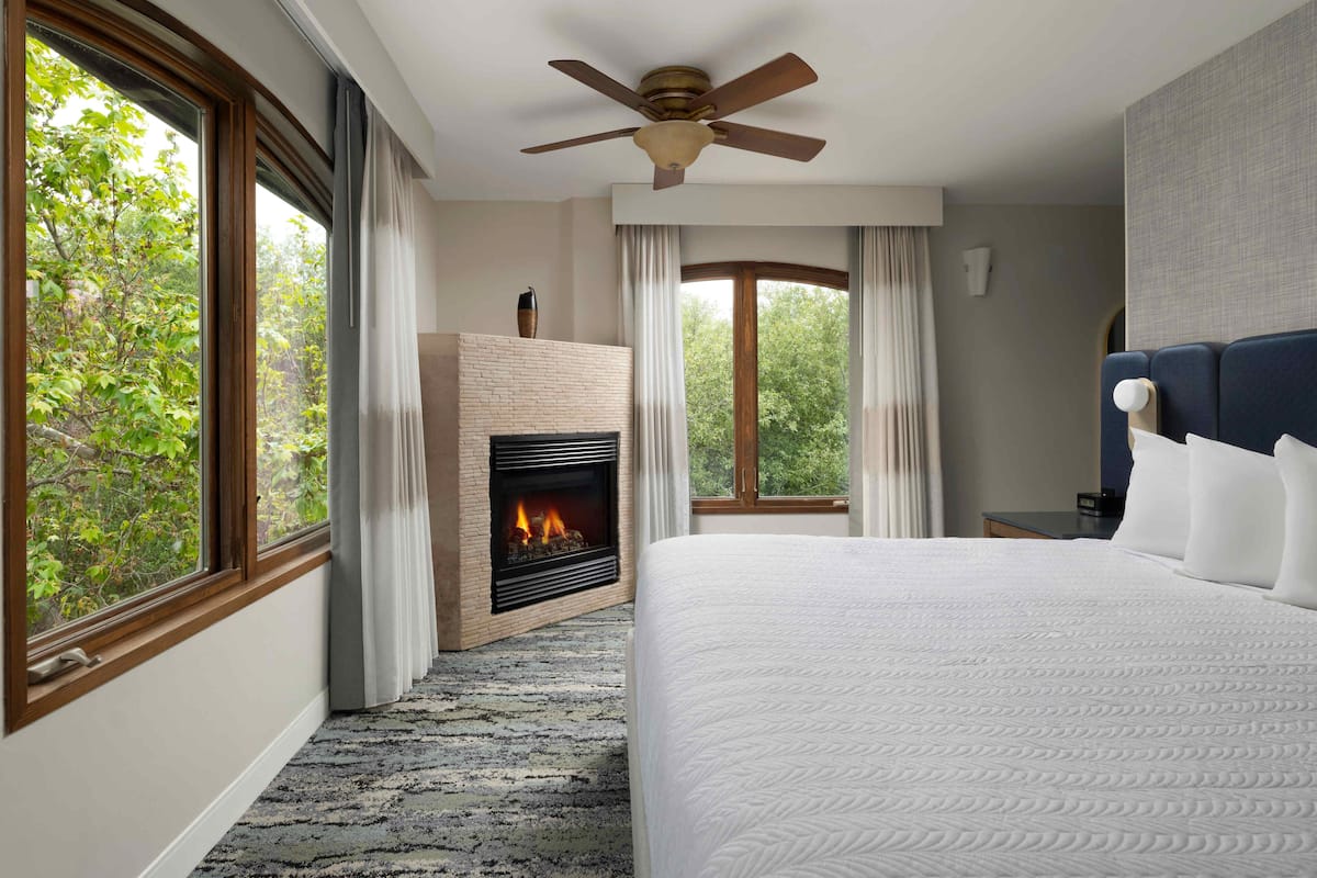 A hotel room with a large bed, a fireplace, a ceiling fan, and a view of trees through the windows.