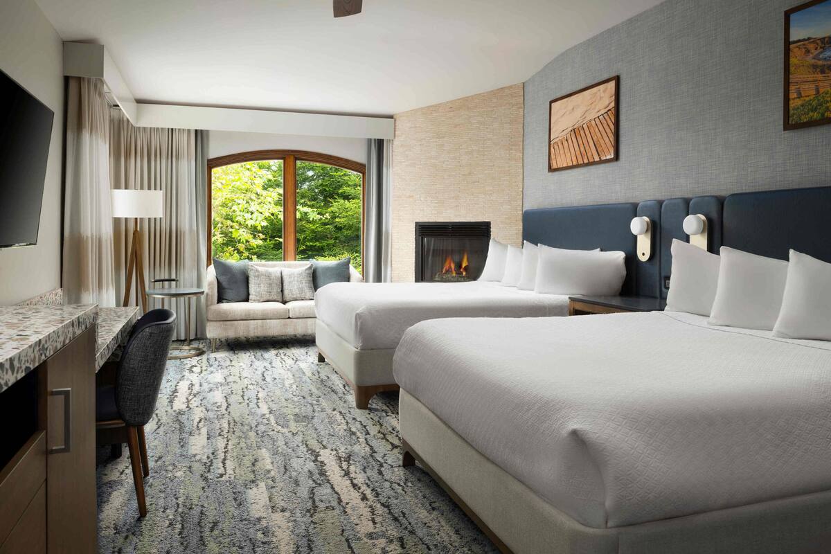 A hotel room with two beds, a desk, a chair, a fireplace, and a large window with a view of greenery.