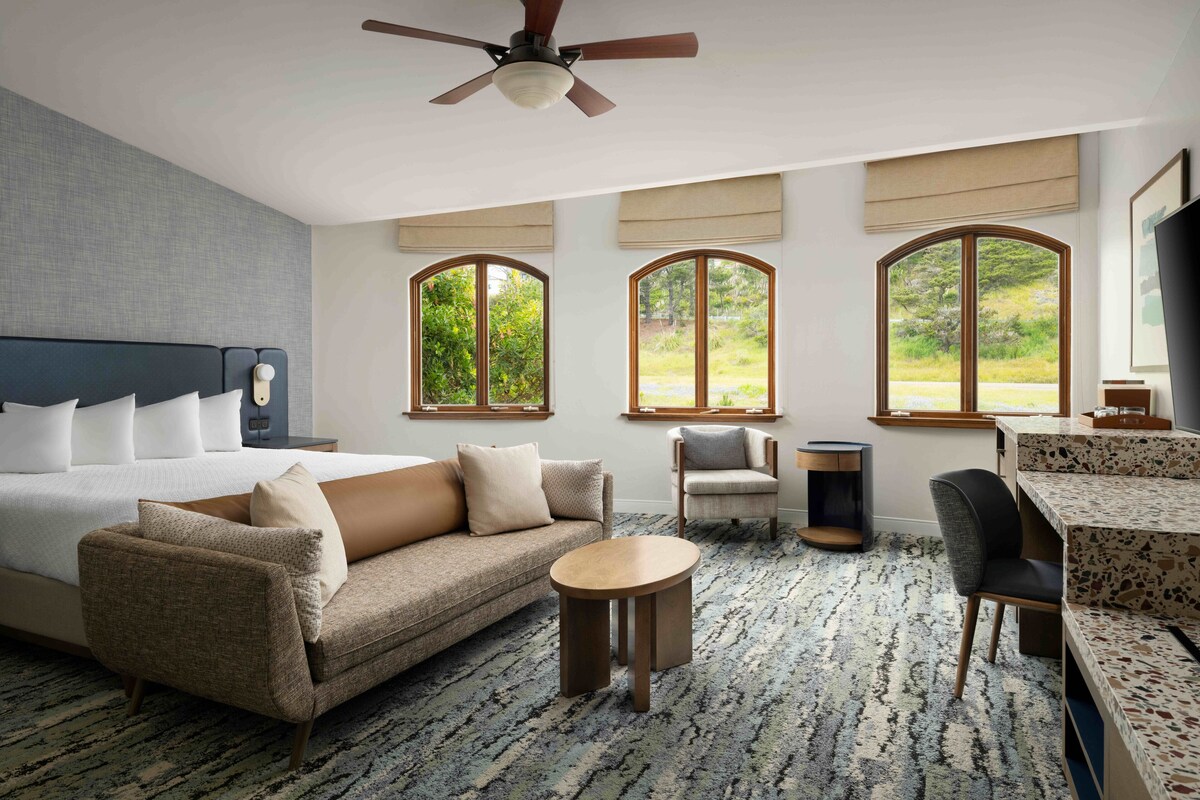 A hotel room with a bed, a sofa, a round table, and a chair. There are large windows with views of greenery.