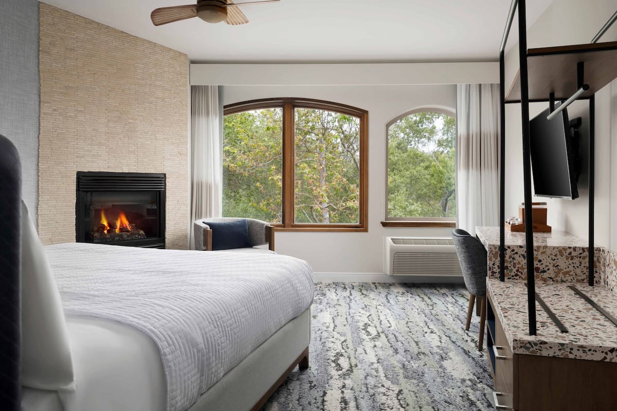 A modern hotel room with a large bed, a fireplace, a view of trees, a desk with a chair, and a television.