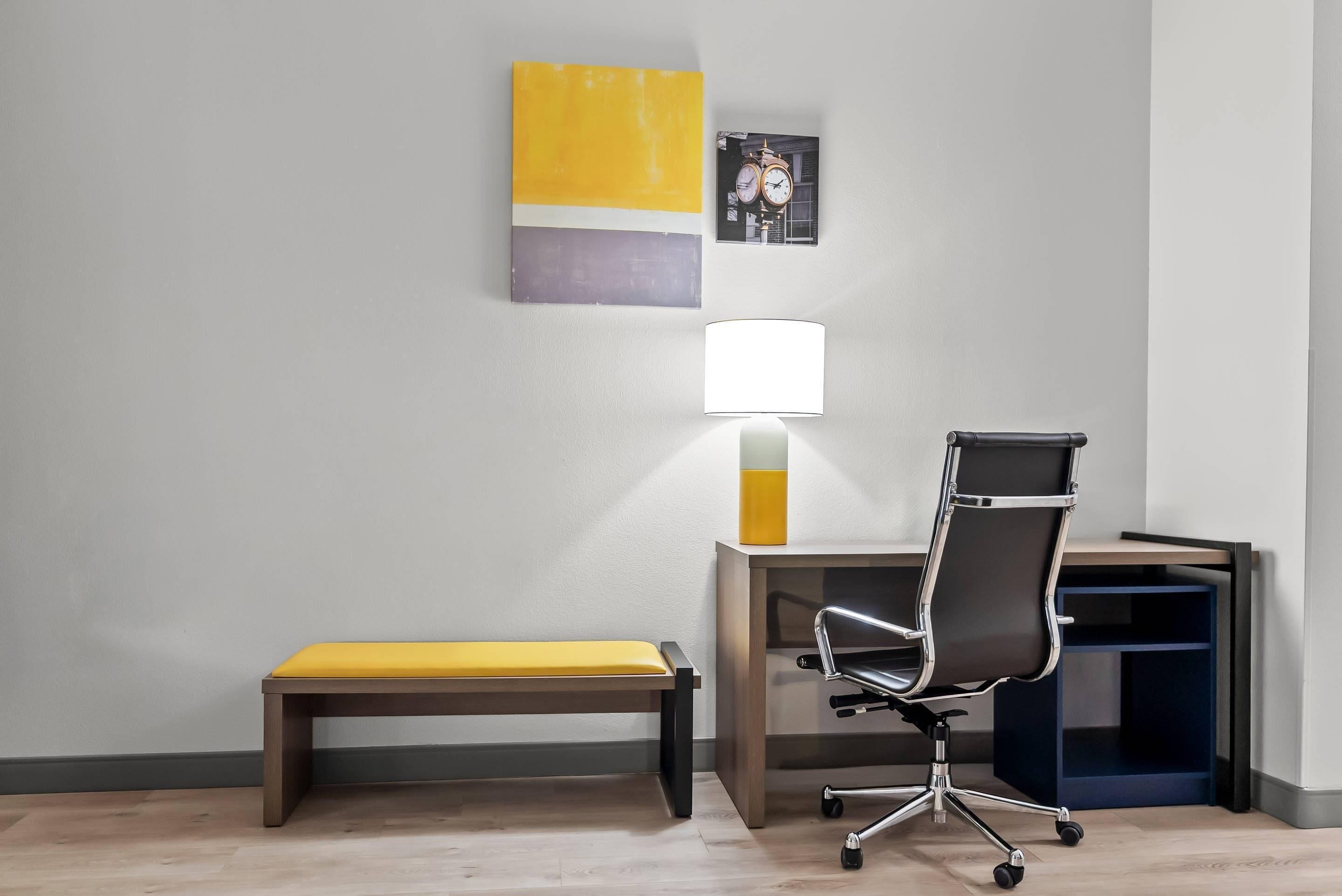 A modern office space with a desk, chair, bench, lamp, and artwork on the wall.