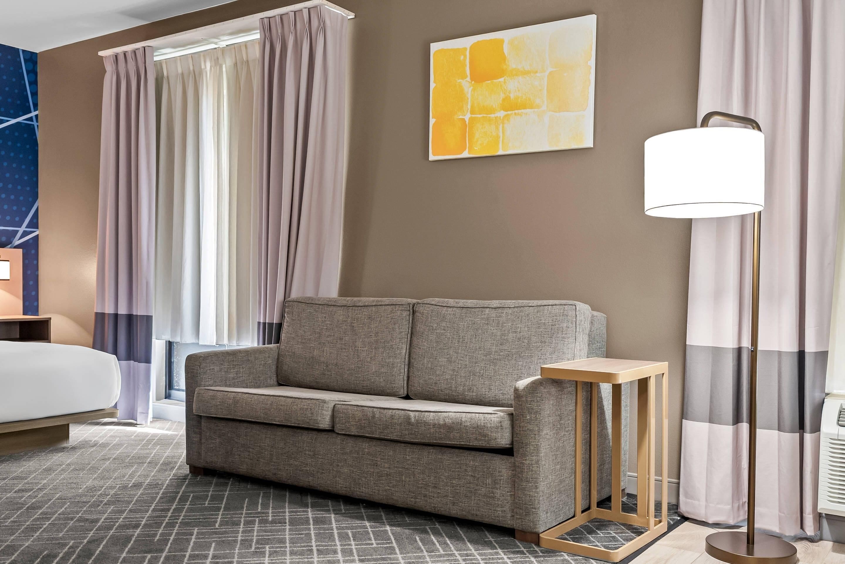 A modern hotel room with a grey sofa, a small wooden side table, a floor lamp, and a large abstract painting on the wall.